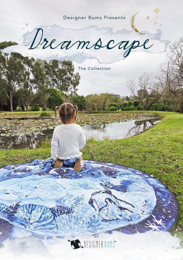 A child sits on a blue and white patterned blanket spread on grass beside a pond in a park, with the text 'Dreamscape The Collection' and 'Designer Bums Presents' overlaid at the top.